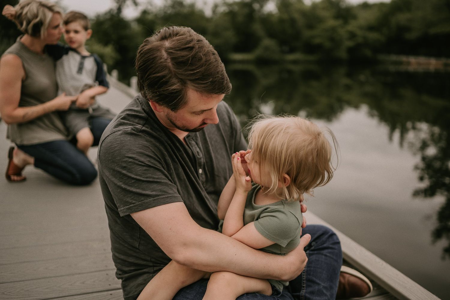 Intimate family moment shared on a wooden dock surrounded by tranquil waters and trees.