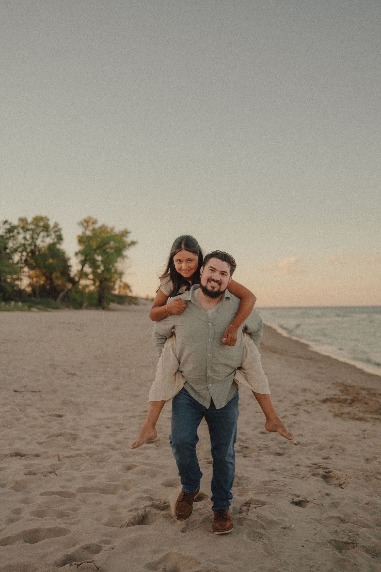Happy father giving daughter a piggyback ride on sandy beach at sunset near ocean shore.
