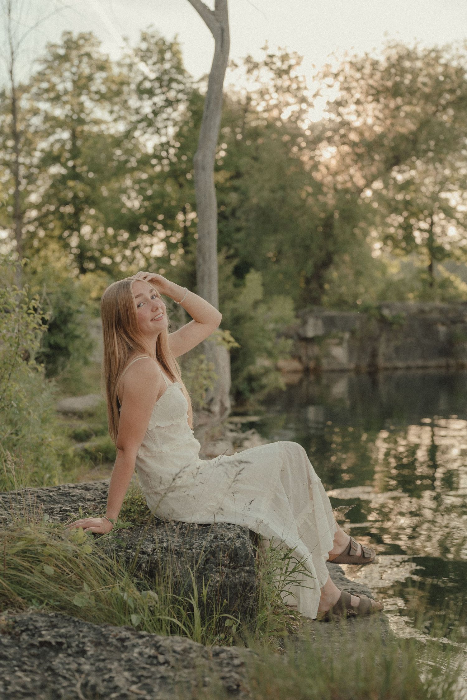 Young woman in white dress sitting on rock by serene pond surrounded by lush green trees at golden hour.