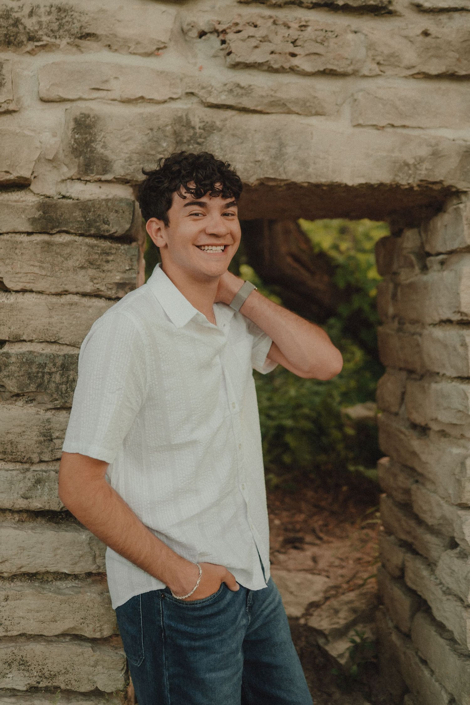 Smiling young man with curly hair in white shirt and jeans leaning against stone wall outdoors.