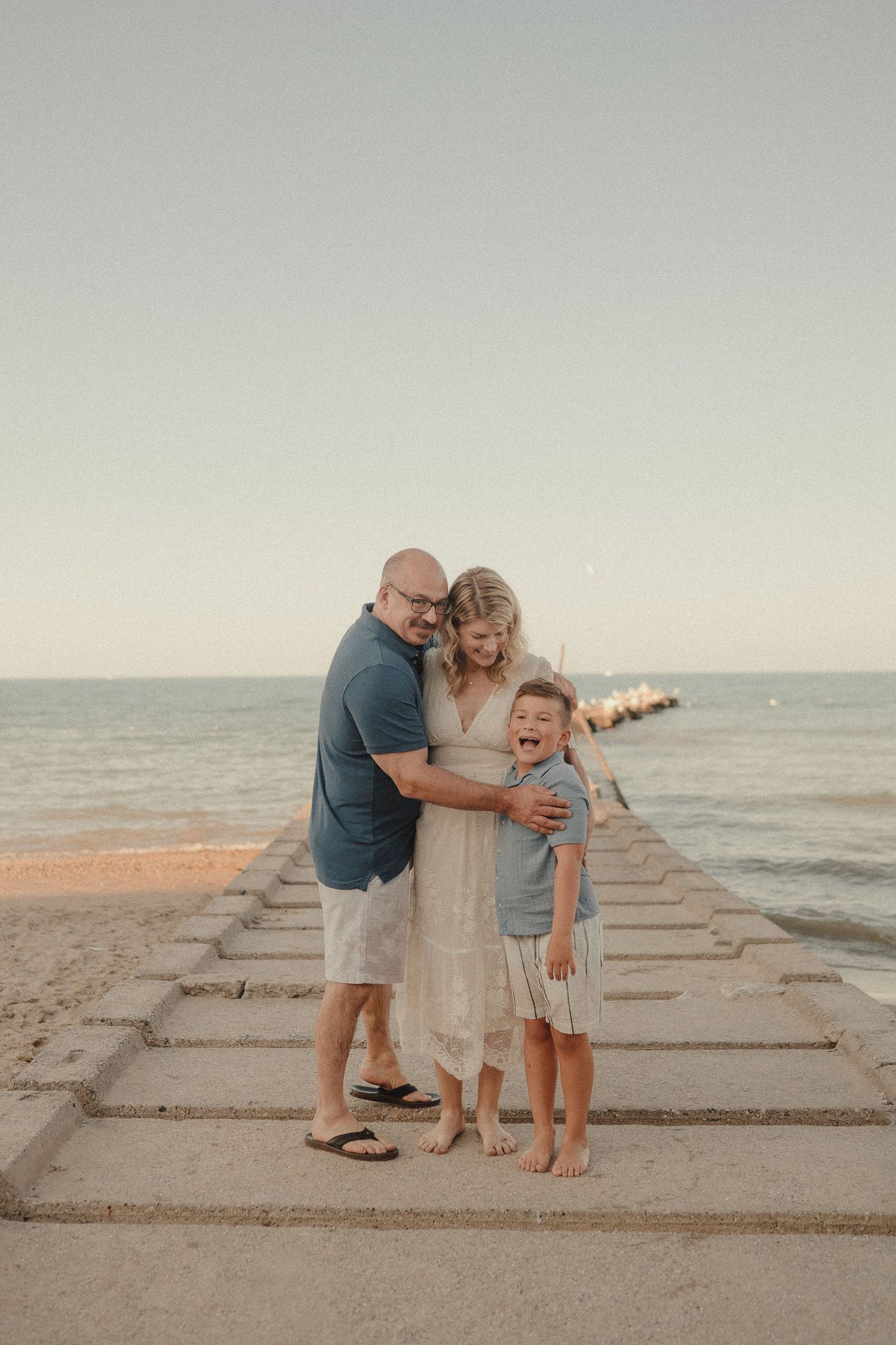 Family of three smiling and hugging on a beach pier at sunset, with calm ocean waves in the background.