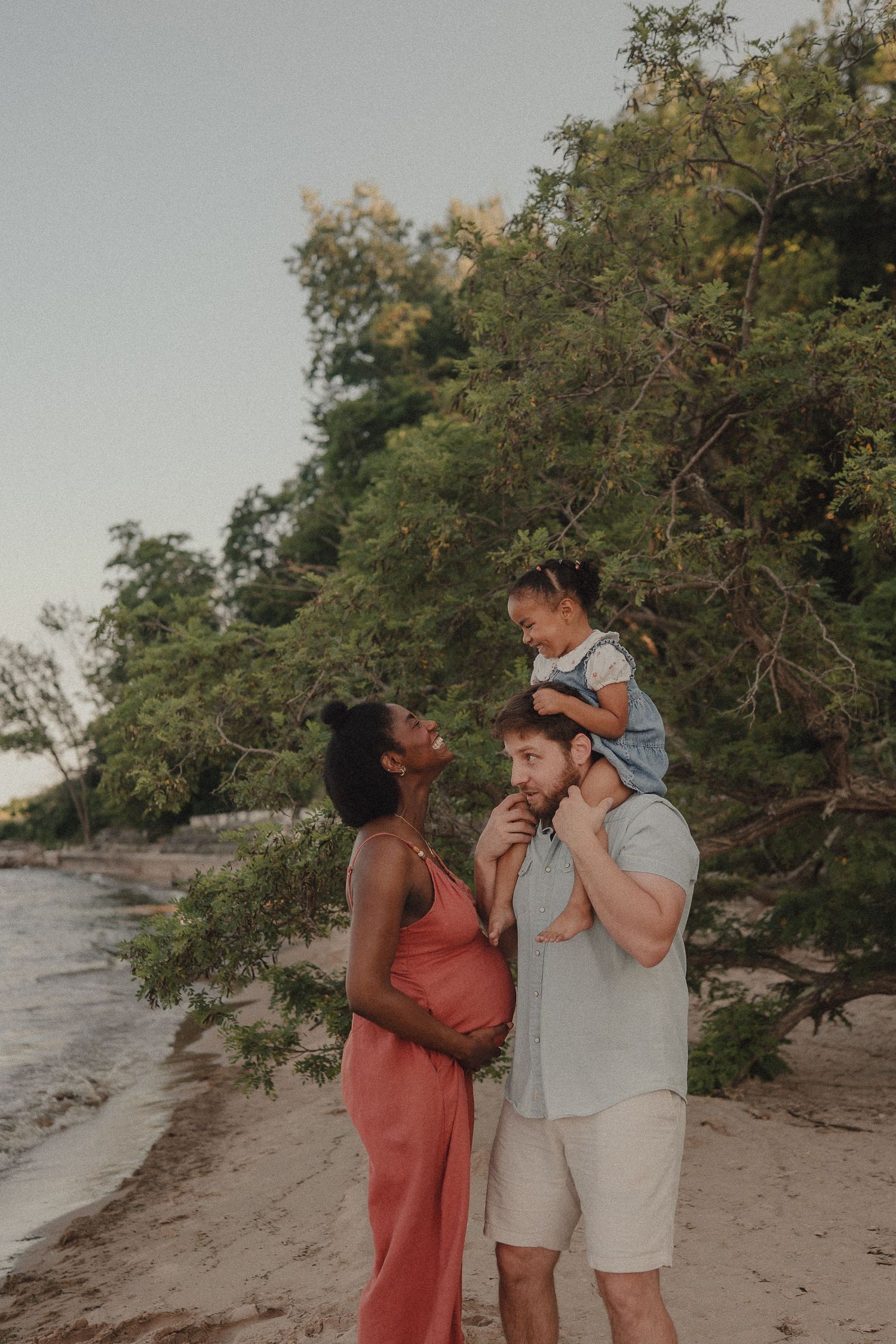 Pregnant woman in pink dress with husband and toddler on shoulders enjoying a beach sunset family photo.