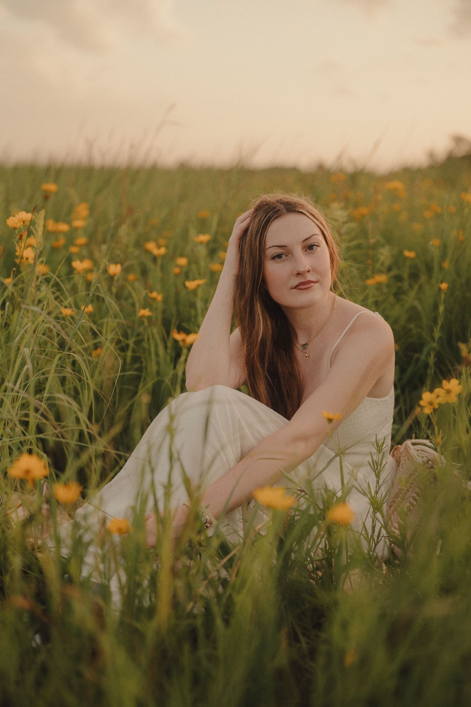 Young woman in white dress sits among yellow wildflowers in a field during golden hour.