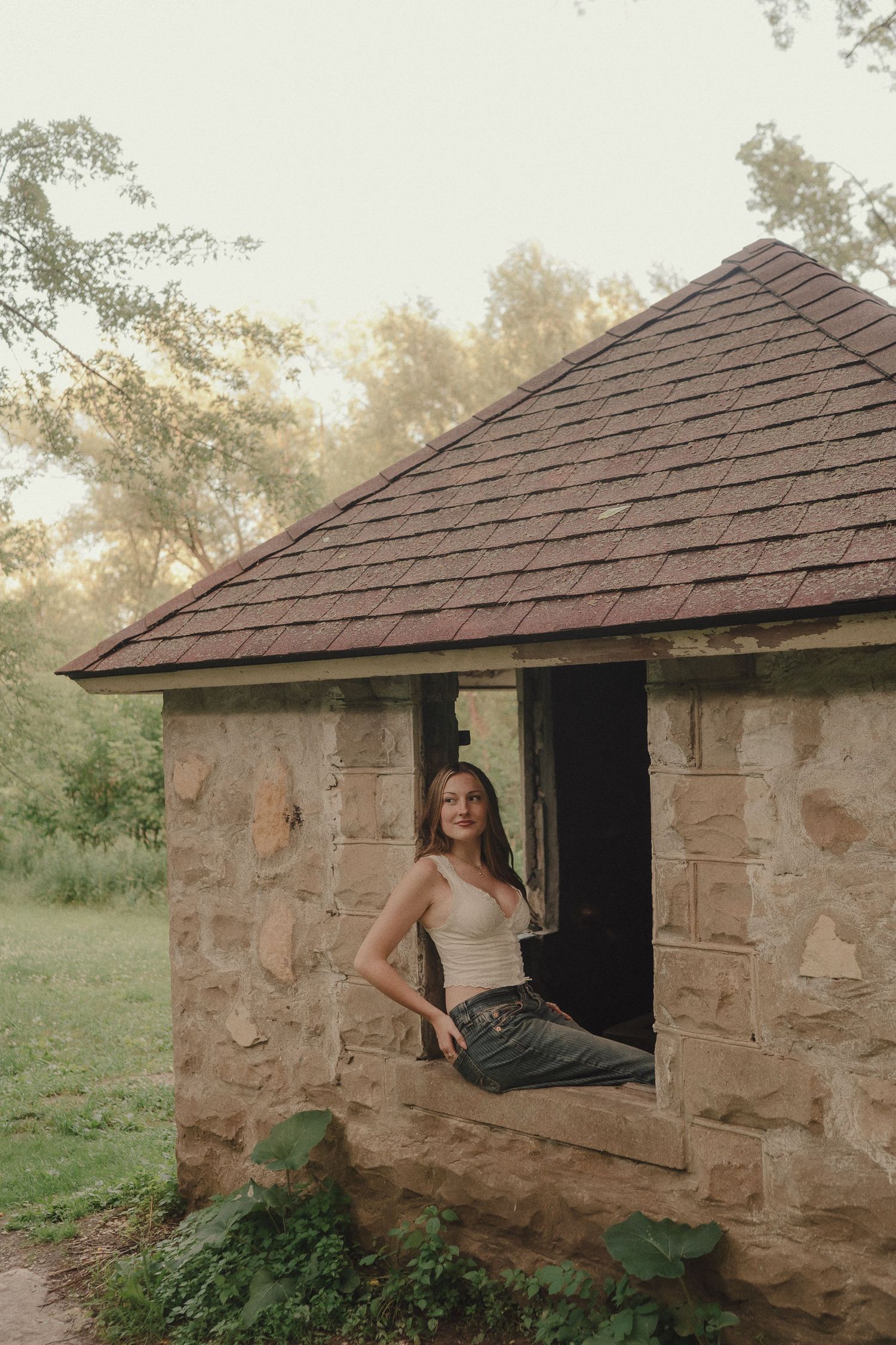 Young woman in white tank top sits in window of rustic stone building at golden hour.
