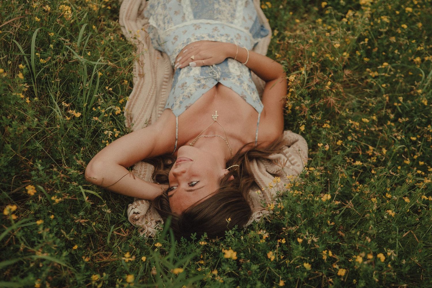 Woman in floral dress lying in a field of yellow wildflowers, smiling peacefully with hands behind her head.