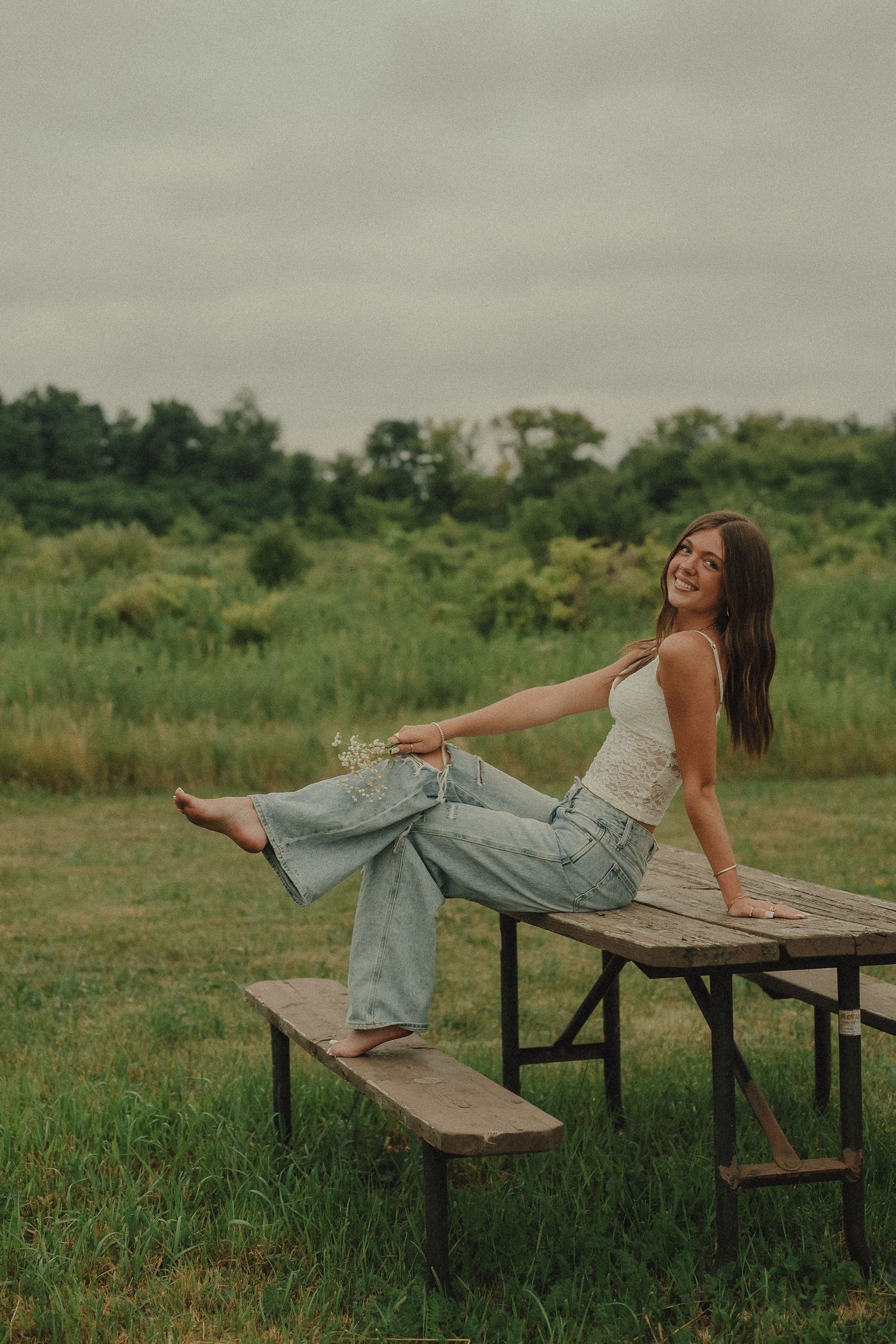 Young woman in white top and wide-leg jeans sitting on wooden picnic table in green meadow.
