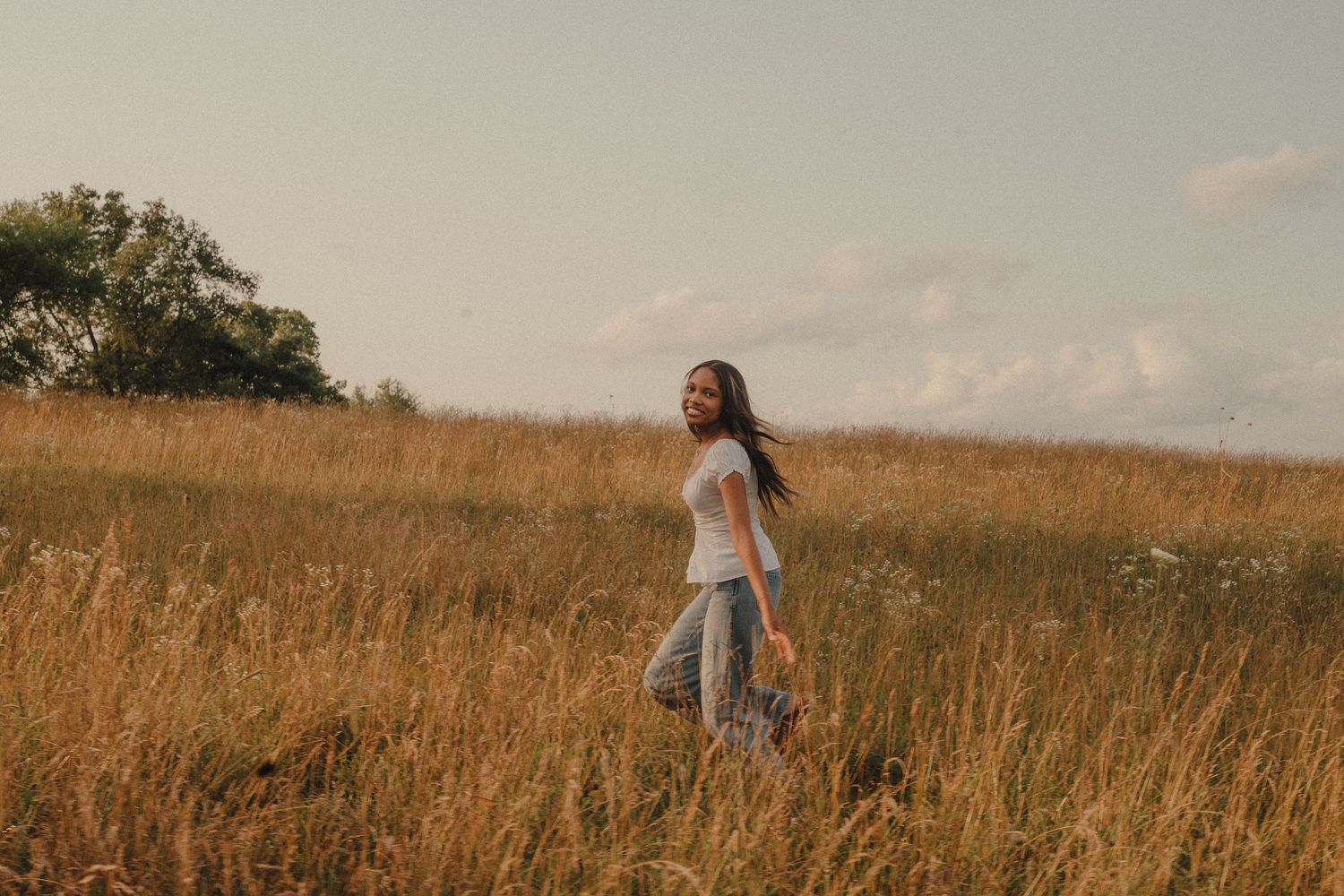 A young woman with long dark hair walks through a golden field of tall grass under a partly cloudy sky.