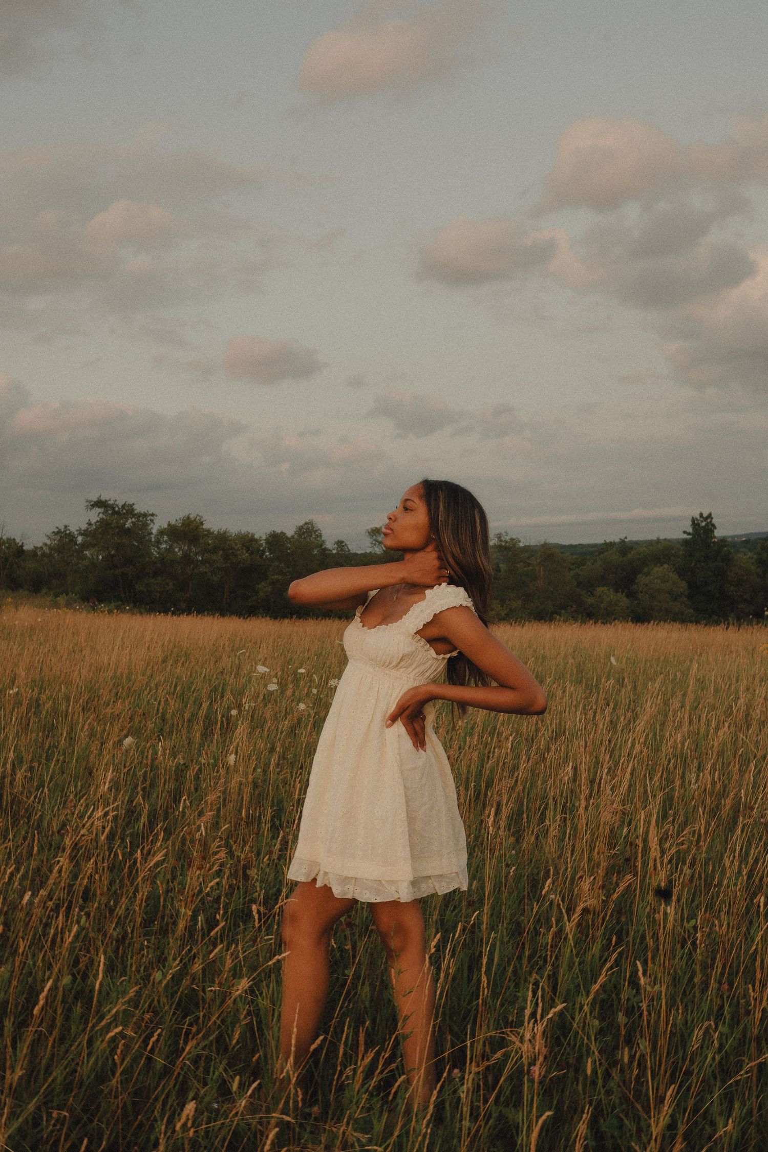 Young woman in white dress posing gracefully in a golden field under a cloudy evening sky.