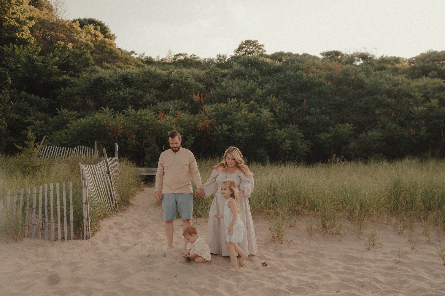 Family of four holding hands walking on sandy beach at dusk.