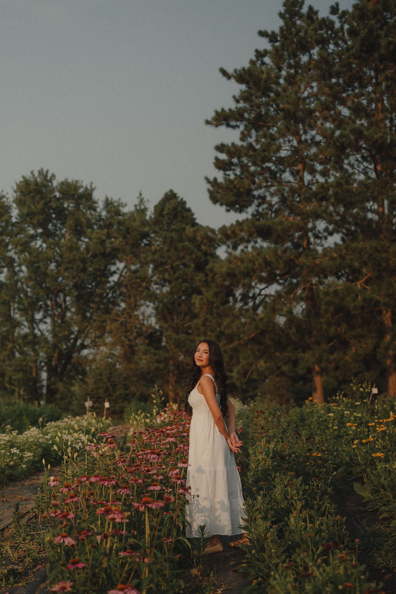Woman in white maxi dress standing in flower garden at dusk with tall trees behind.