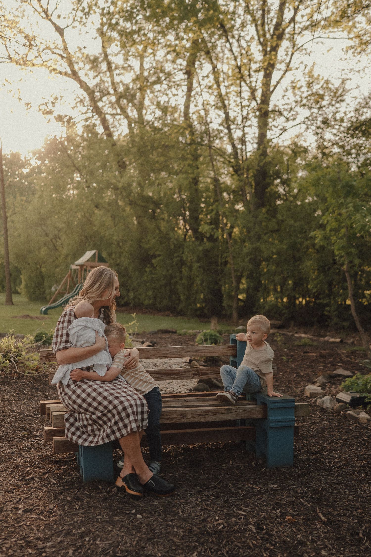Mother in plaid dress sitting on bench holding newborn while young toddler plays beside her outdoors.