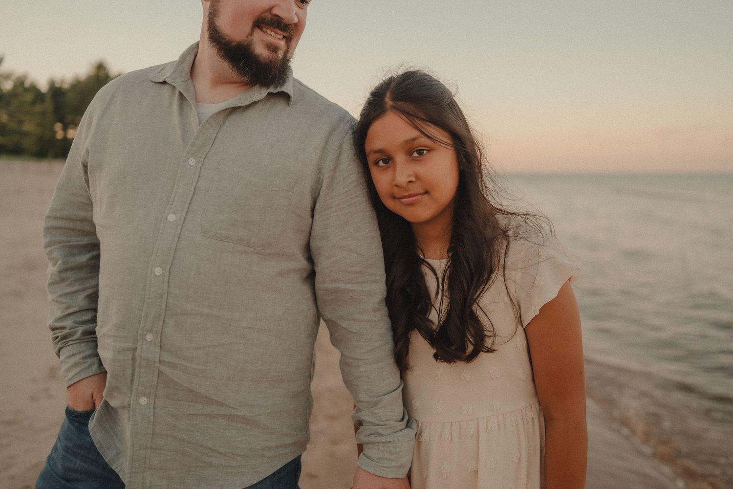 A man and young girl hold hands and pose together on a beach during a warm golden sunset.