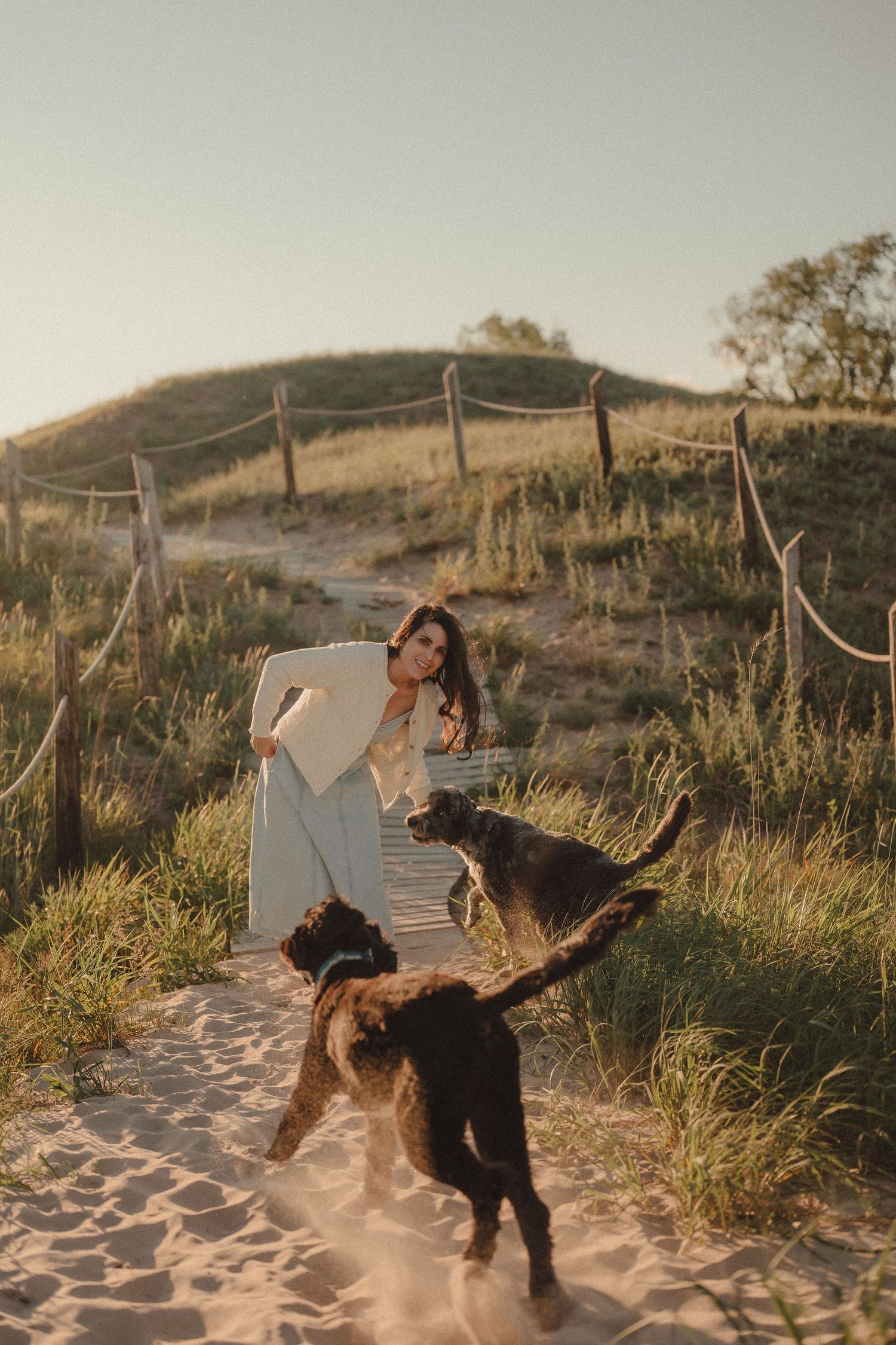 Woman in white dress plays with two dogs on a sandy beach path at golden hour.