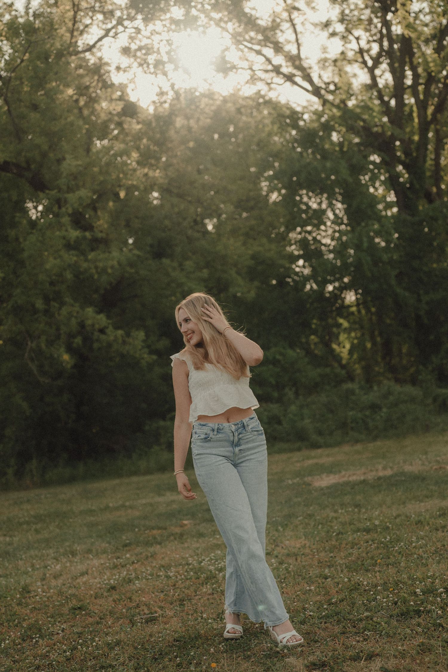 Young woman in white crop top and light wash flare jeans standing in a sunlit green field at golden hour.