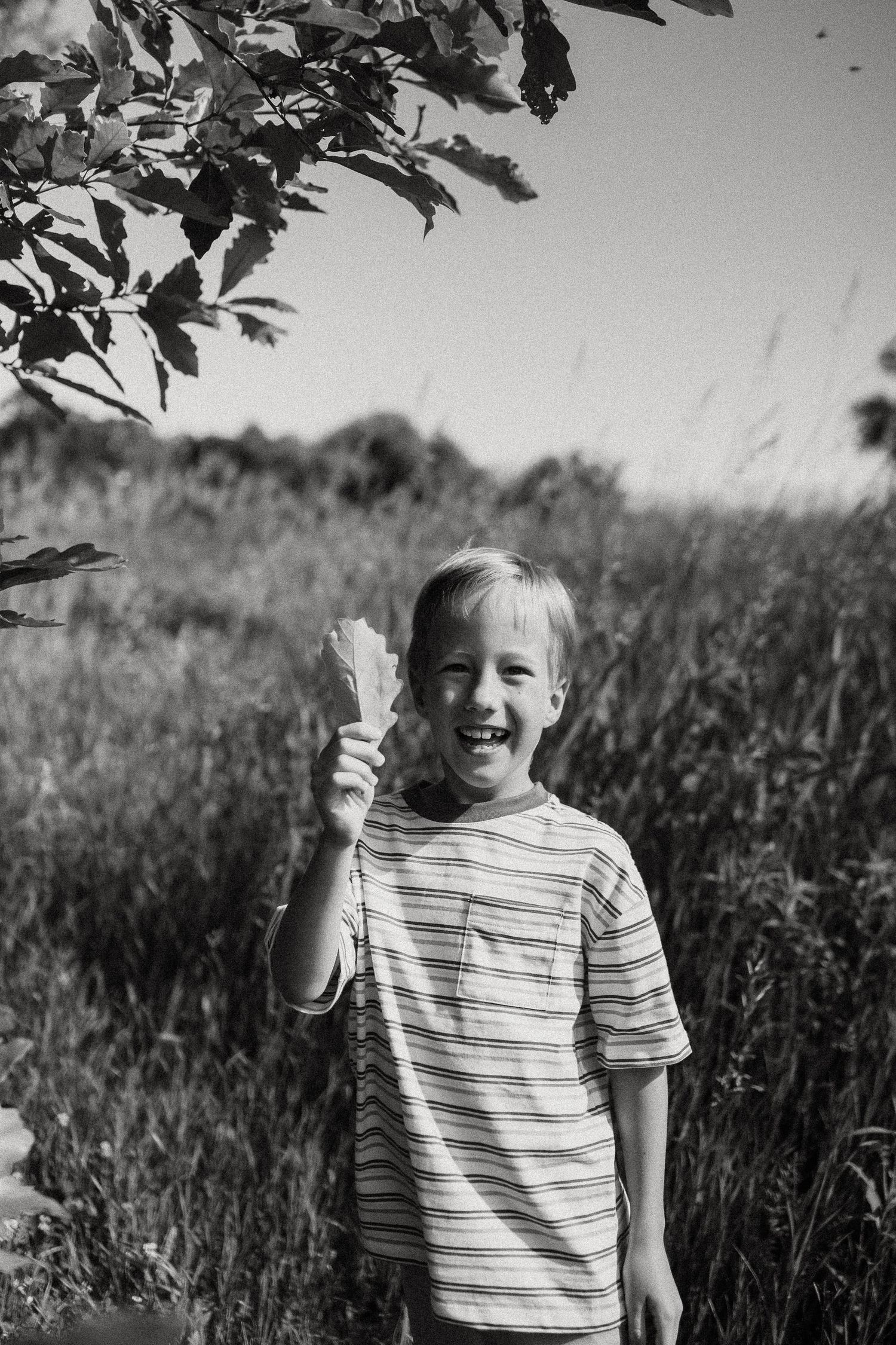 A joyful blonde boy holds a leaf while laughing outdoors in a black and white photo.