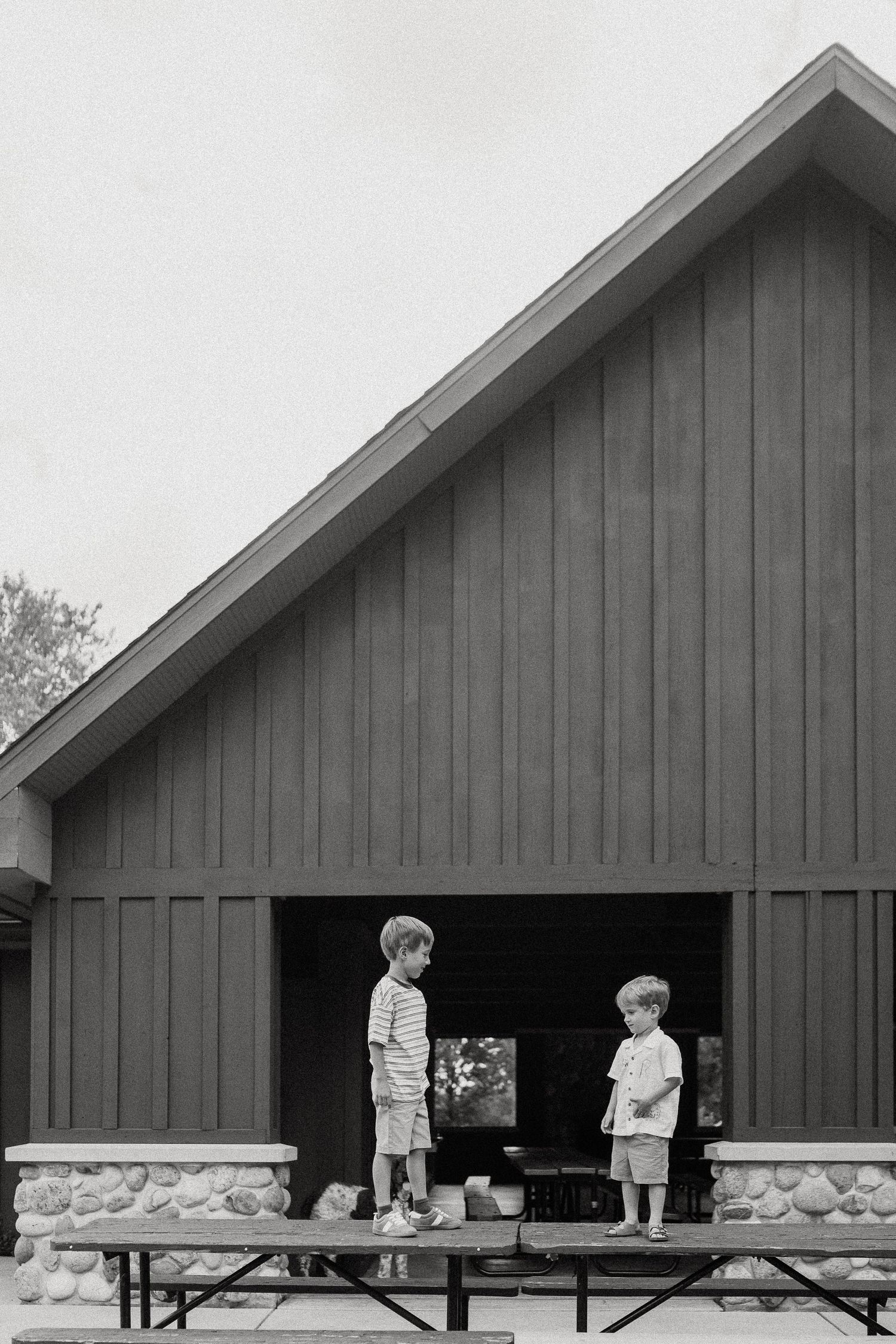 Two young boys standing on a picnic table bench outside a rustic barn-style building in a black and white photo.