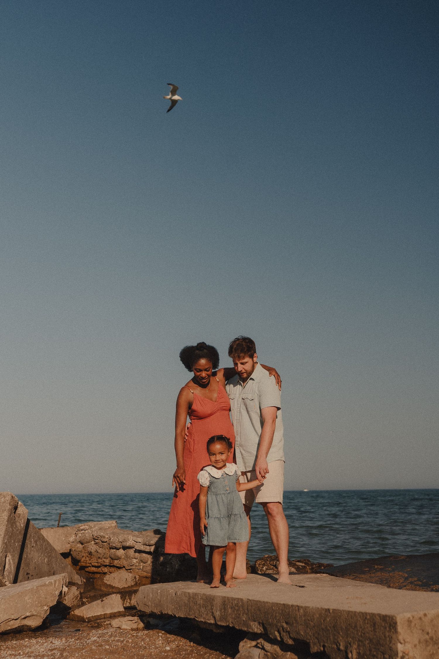 An interracial family of three stands on rocky shoreline with a seagull flying overhead.