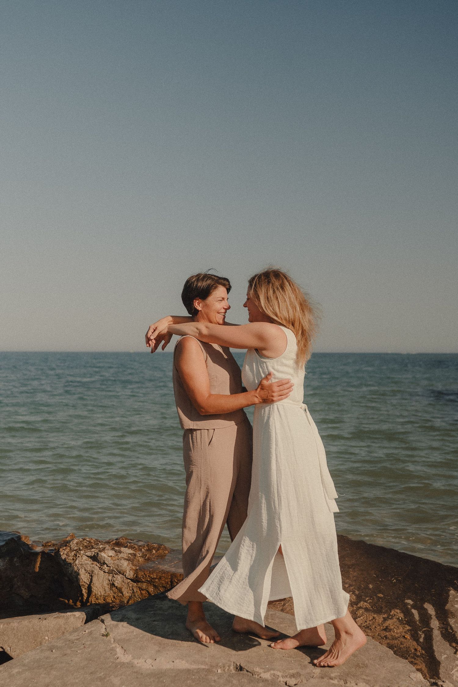 Two women embrace and smile at each other while standing barefoot on rocky shore with calm ocean backdrop.