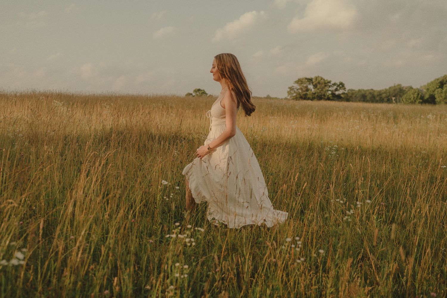 A woman in a lace white dress walks through tall golden grass in a serene countryside field.