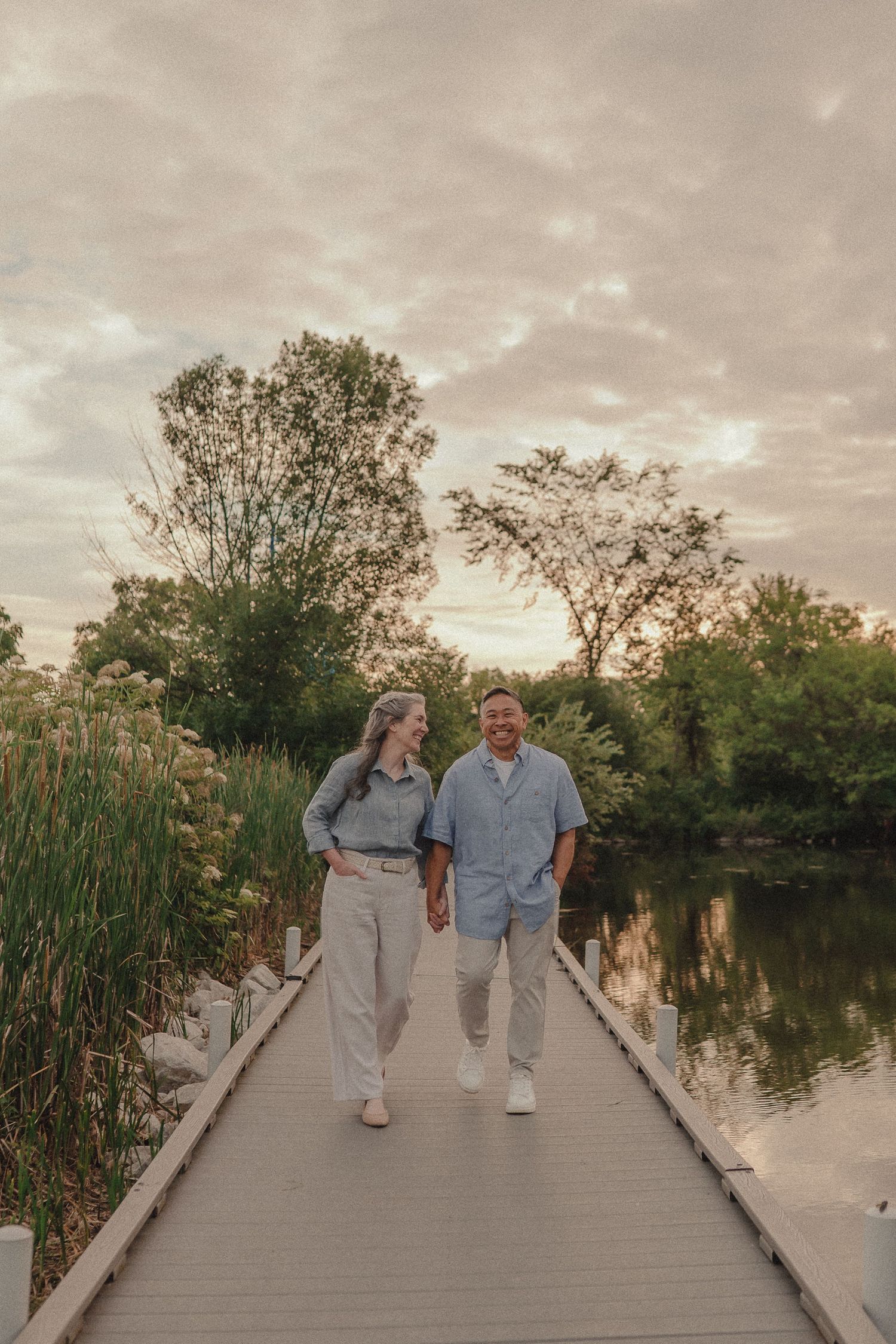 Couple holding hands walking on a wooden dock by a serene lake surrounded by lush green trees at sunset.