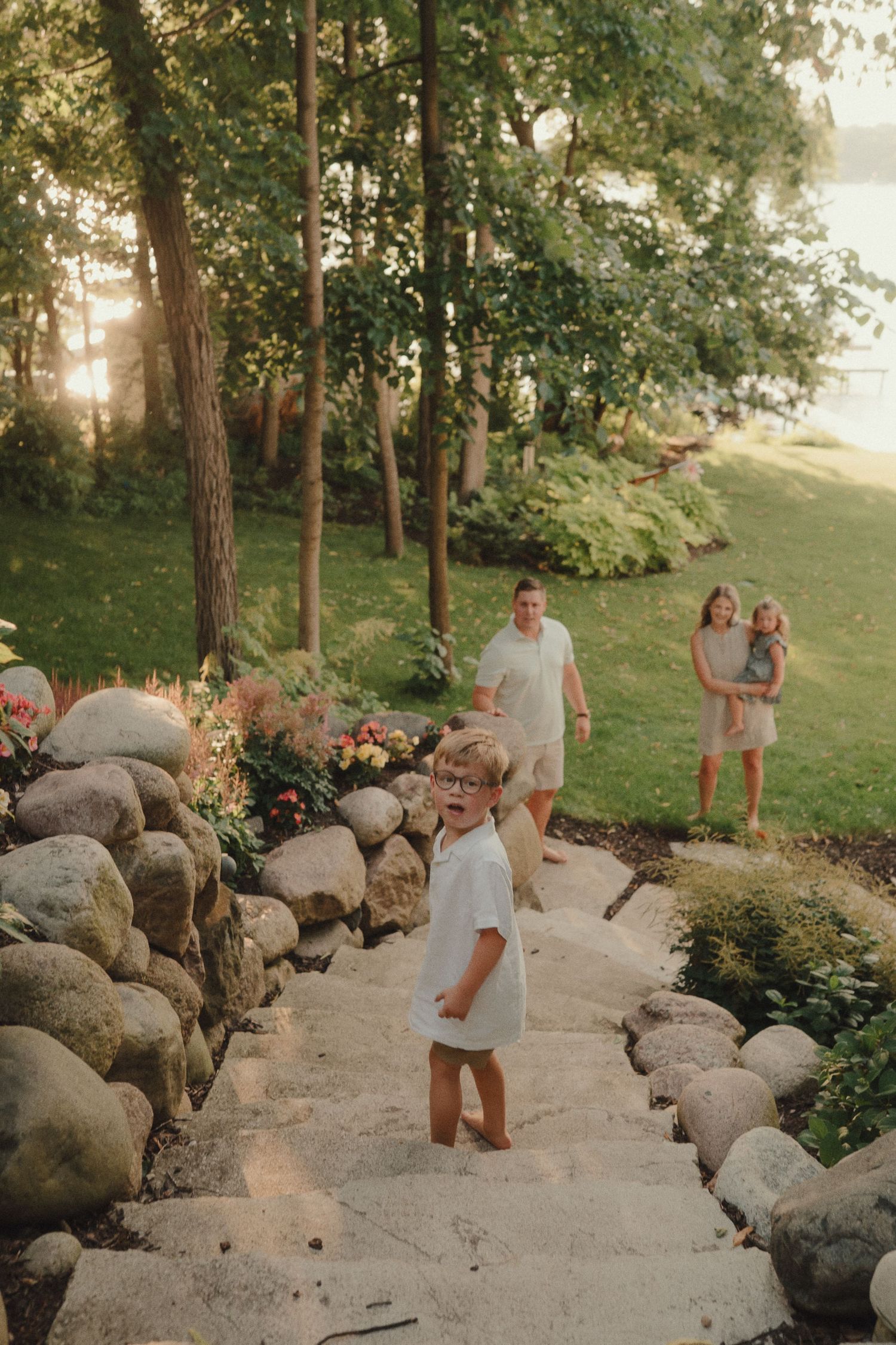 Family walking down stone garden steps at golden hour near a lake, surrounded by lush greenery and flower beds.