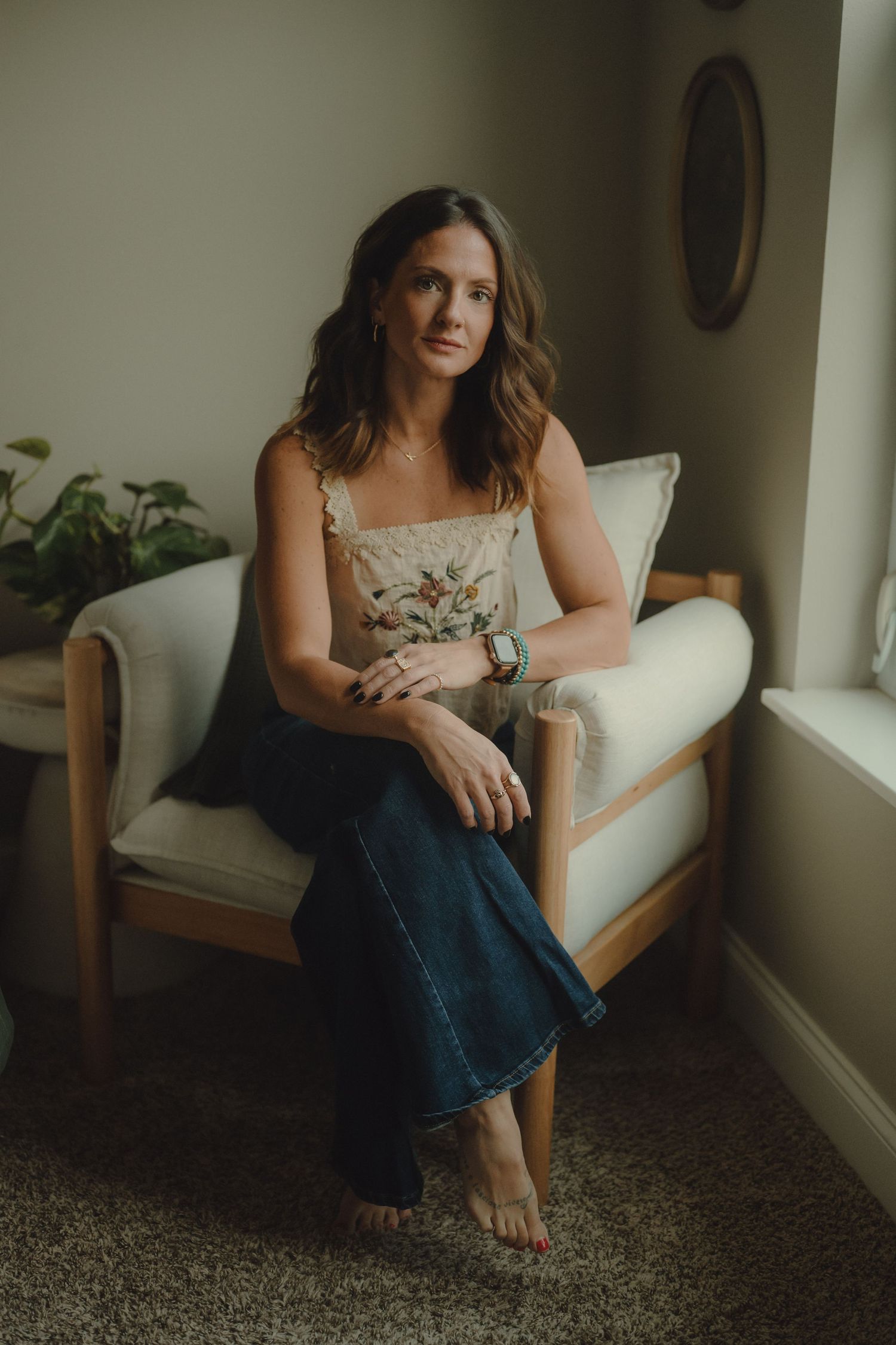 Woman with wavy brown hair sits barefoot in a cozy cushioned wooden armchair wearing a floral cami and wide-leg jeans near a window.