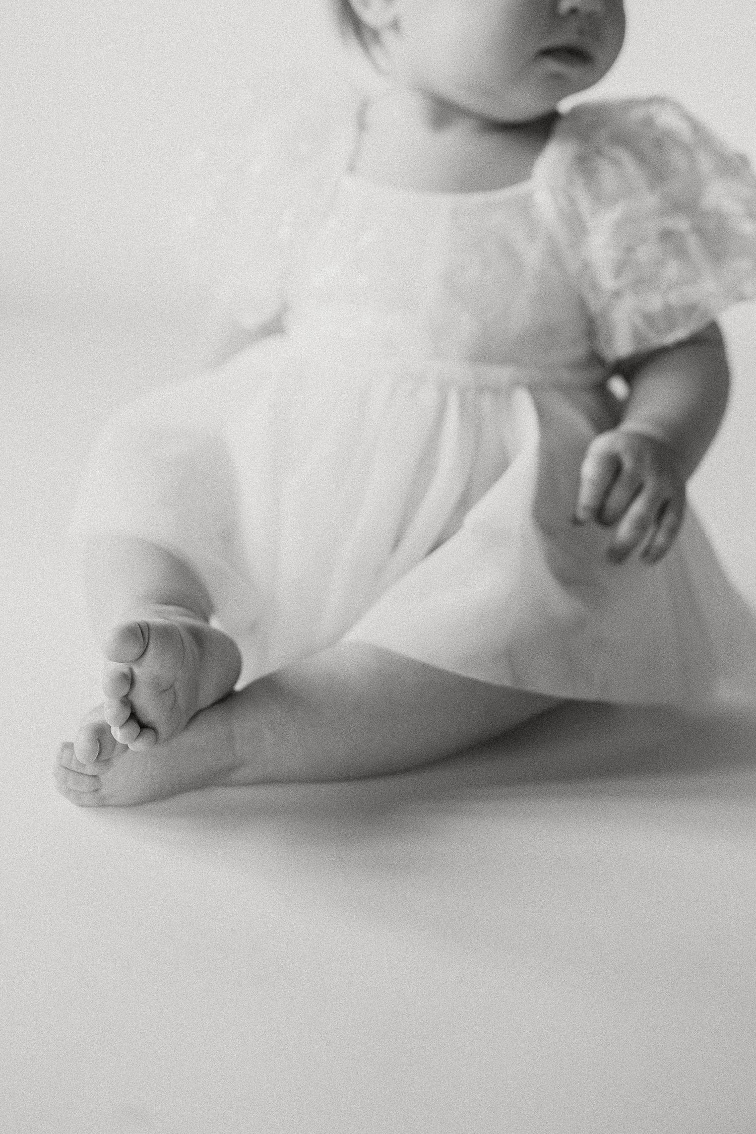 Black and white close-up of a baby in a white lace dress sitting with bare feet extended.