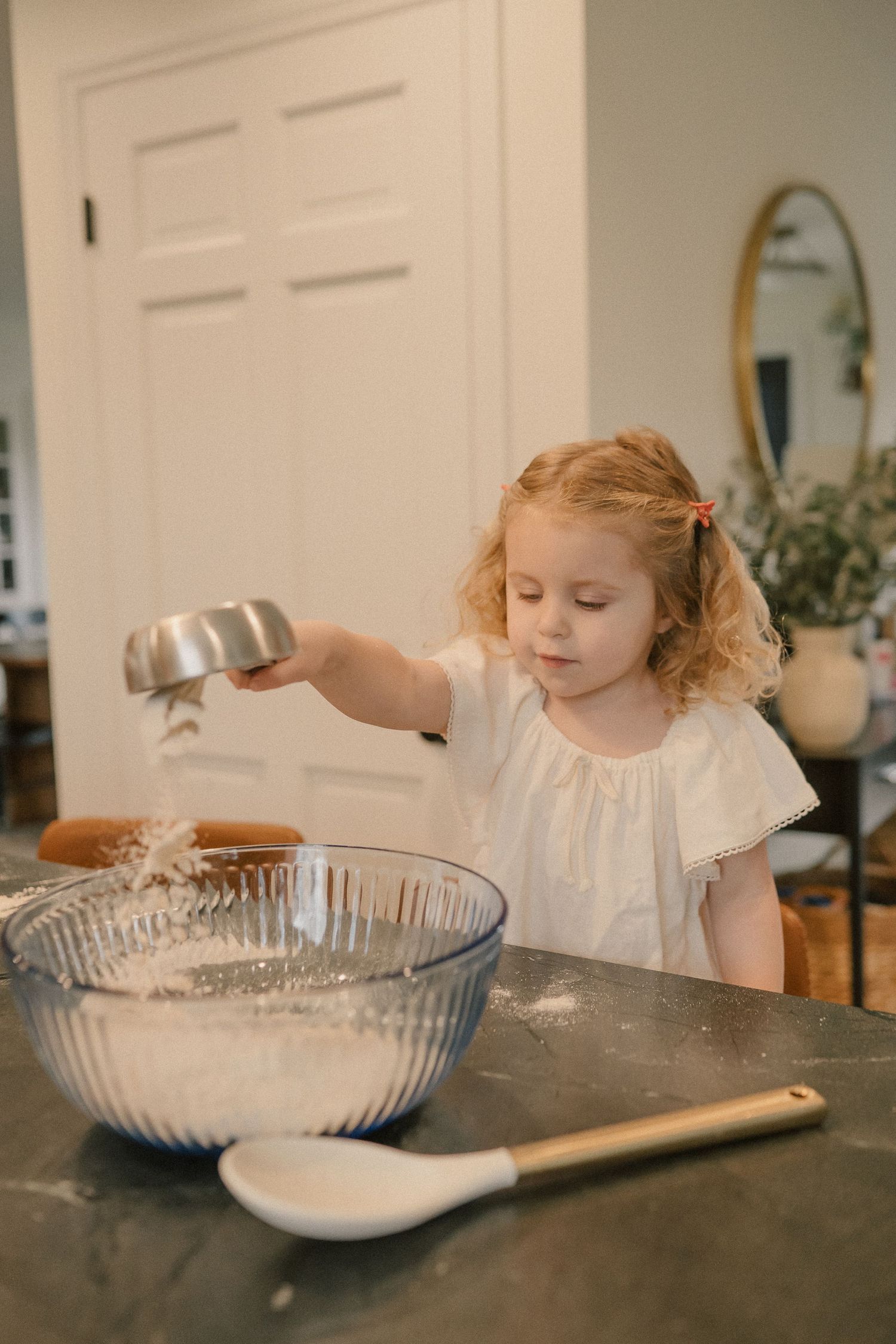 A young girl with curly hair pours flour into a glass mixing bowl on a dark countertop.