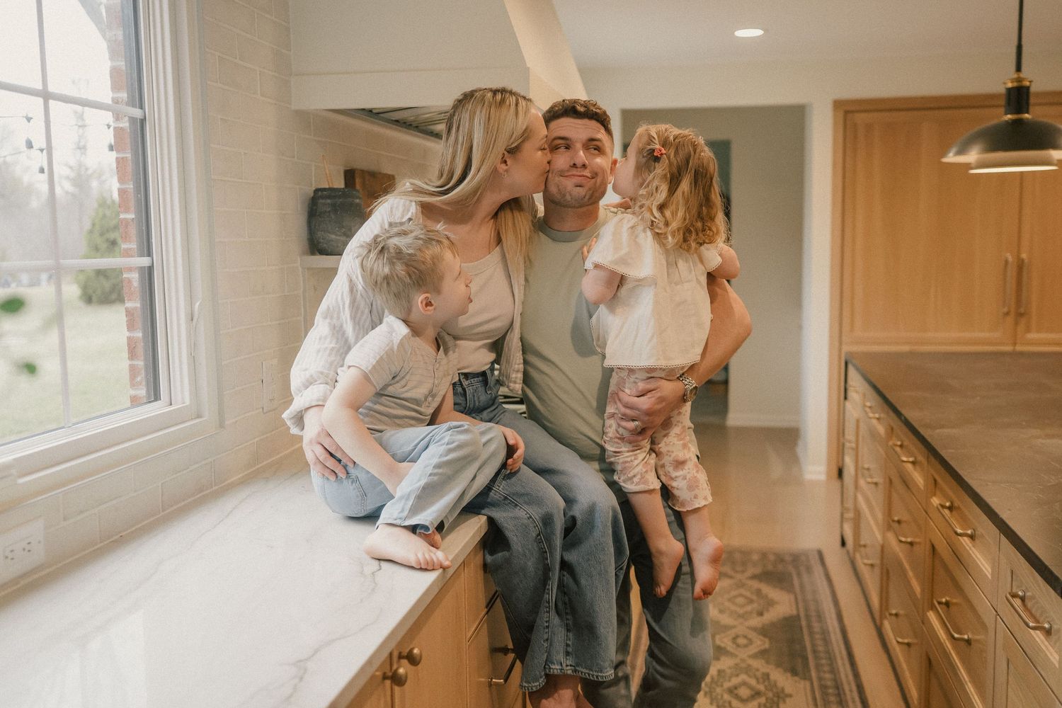 A smiling family of four shares a warm embrace in a bright modern kitchen.