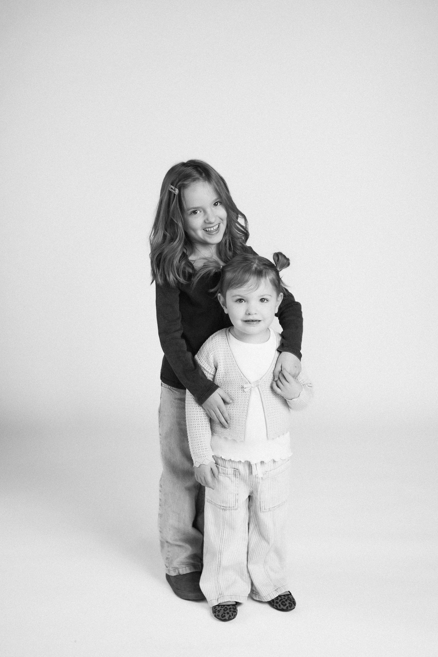 Black and white photo series showing a toddler in white overalls being supported while learning to stand and walk.