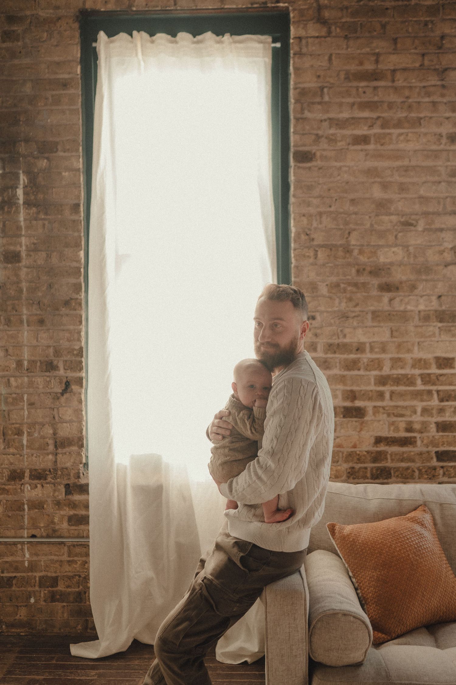 A tender moment between parent and child by a tall window with exposed brick walls and sheer curtains.
