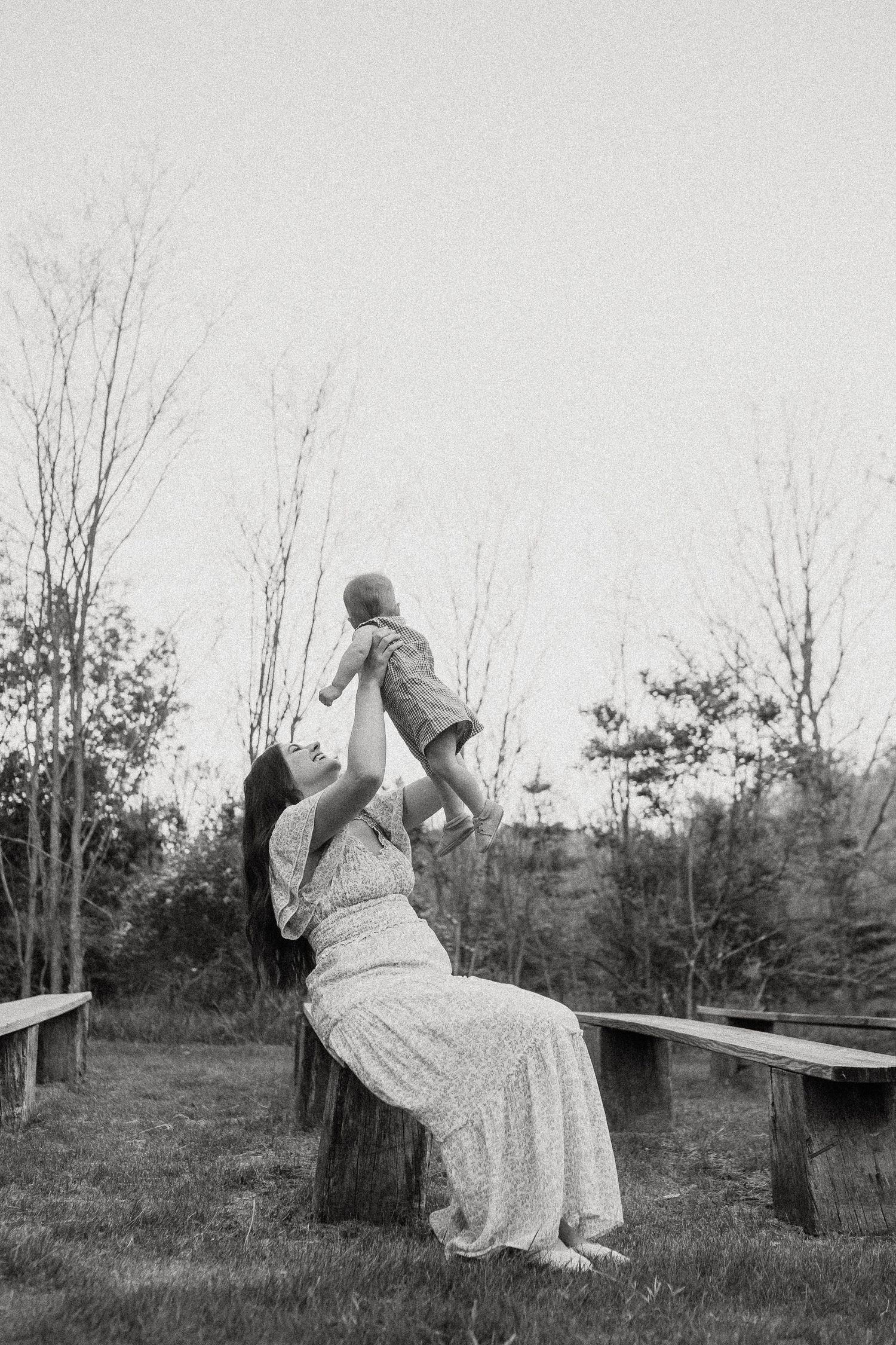 Black and white photo of mother sitting on stump lifting baby joyfully into the air outdoors.