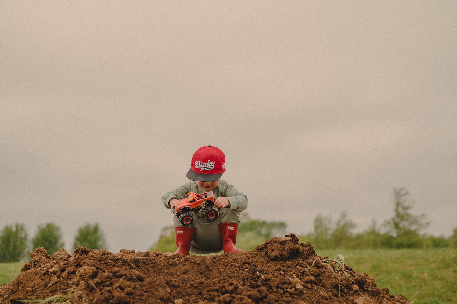 Toddler in red cap and rain boots plays with toy truck atop a large dirt mound outdoors.