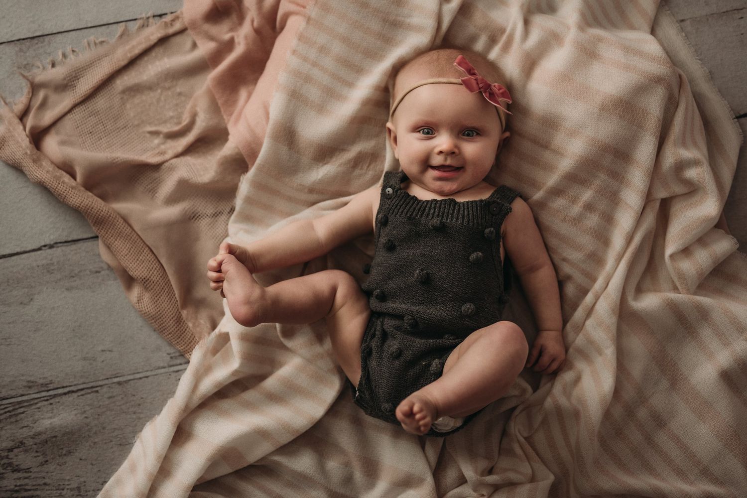 Baby in dark romper and pink headband lying on peach colored blanket.