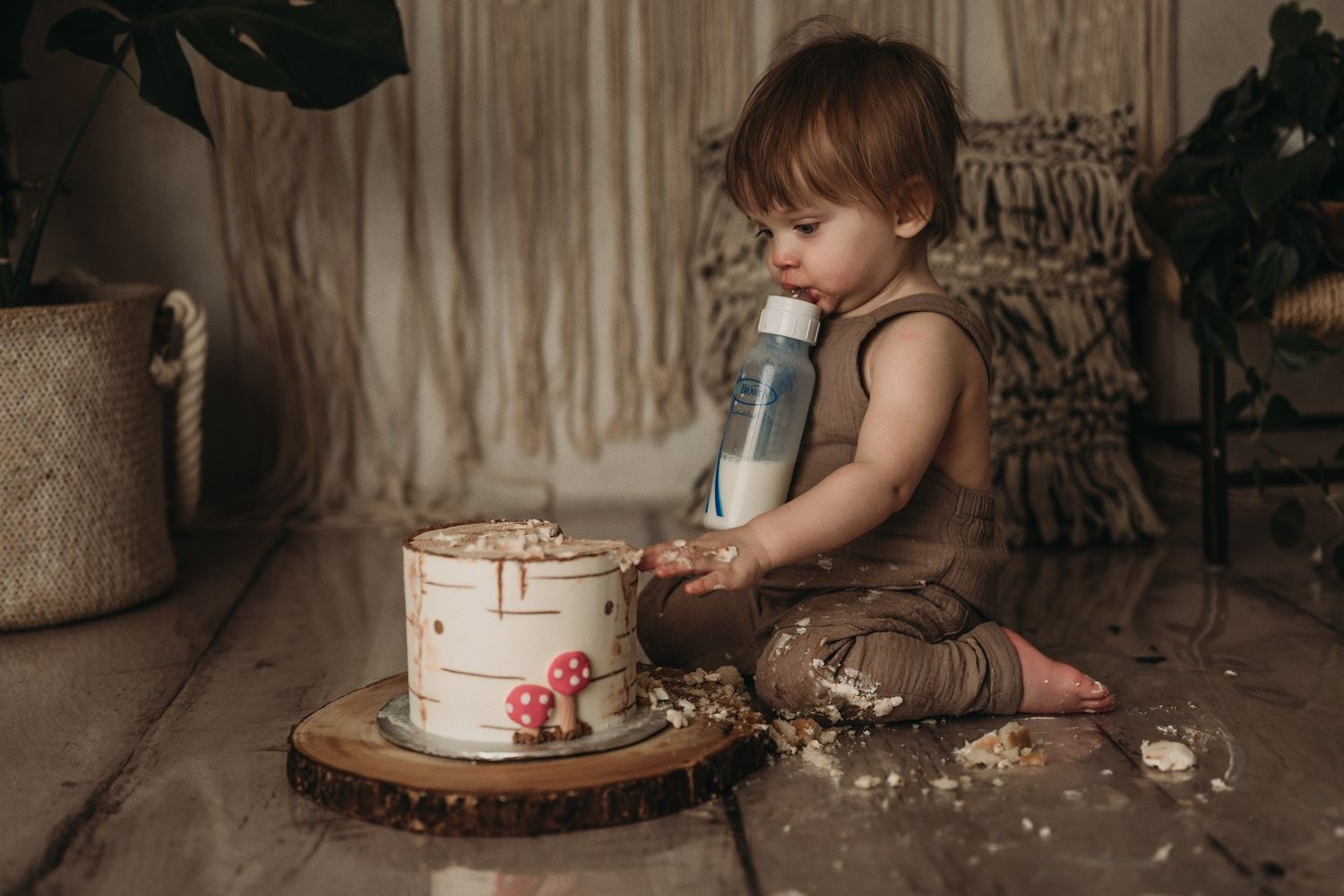 Small toddler in brown outfit sitting on floor with white cake on wooden board during cake smash photoshoot.