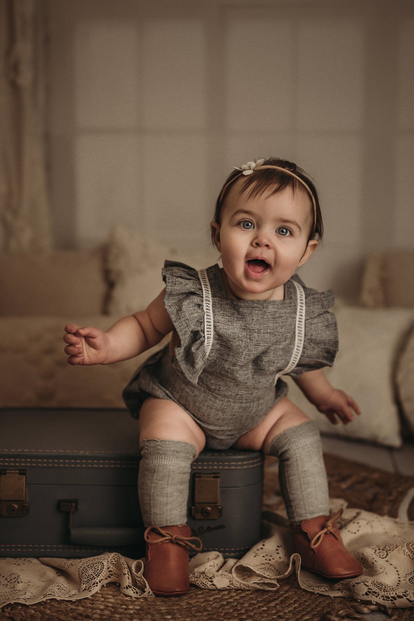 Baby in grey romper with ruffles and knee socks poses excitedly on vintage suitcase.