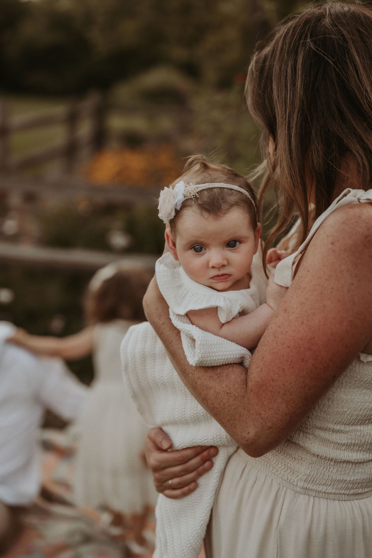 A baby in a white dress and headband is held in someone's arms outdoors.