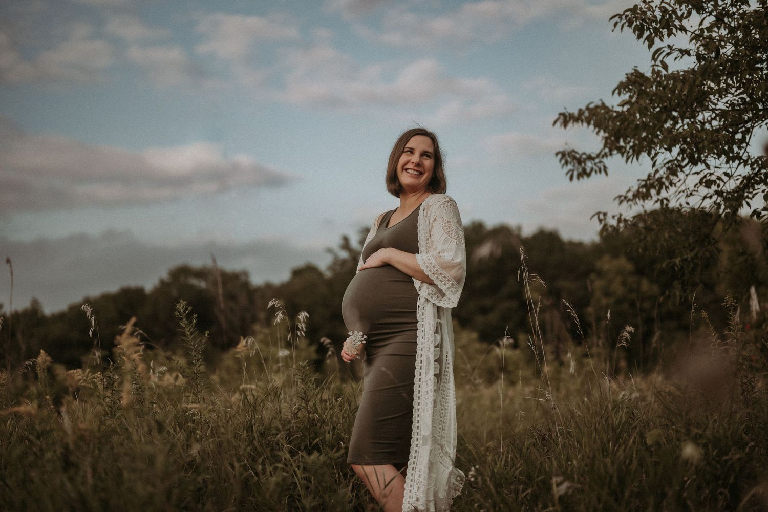An expectant mother poses in a brown dress and white lace cardigan in a meadow.
