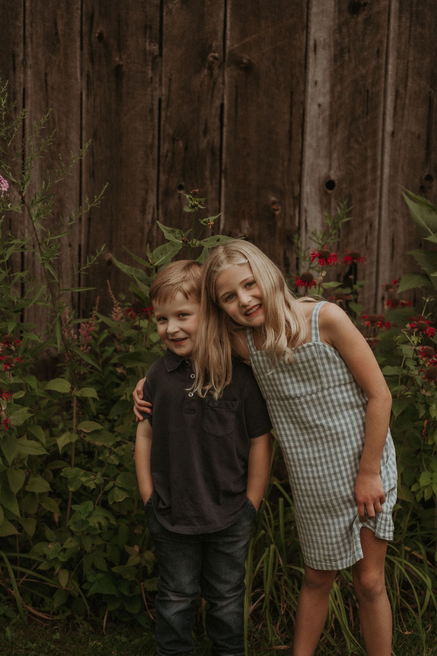 Two children share a playful moment against weathered wooden wall with blooming flowers.