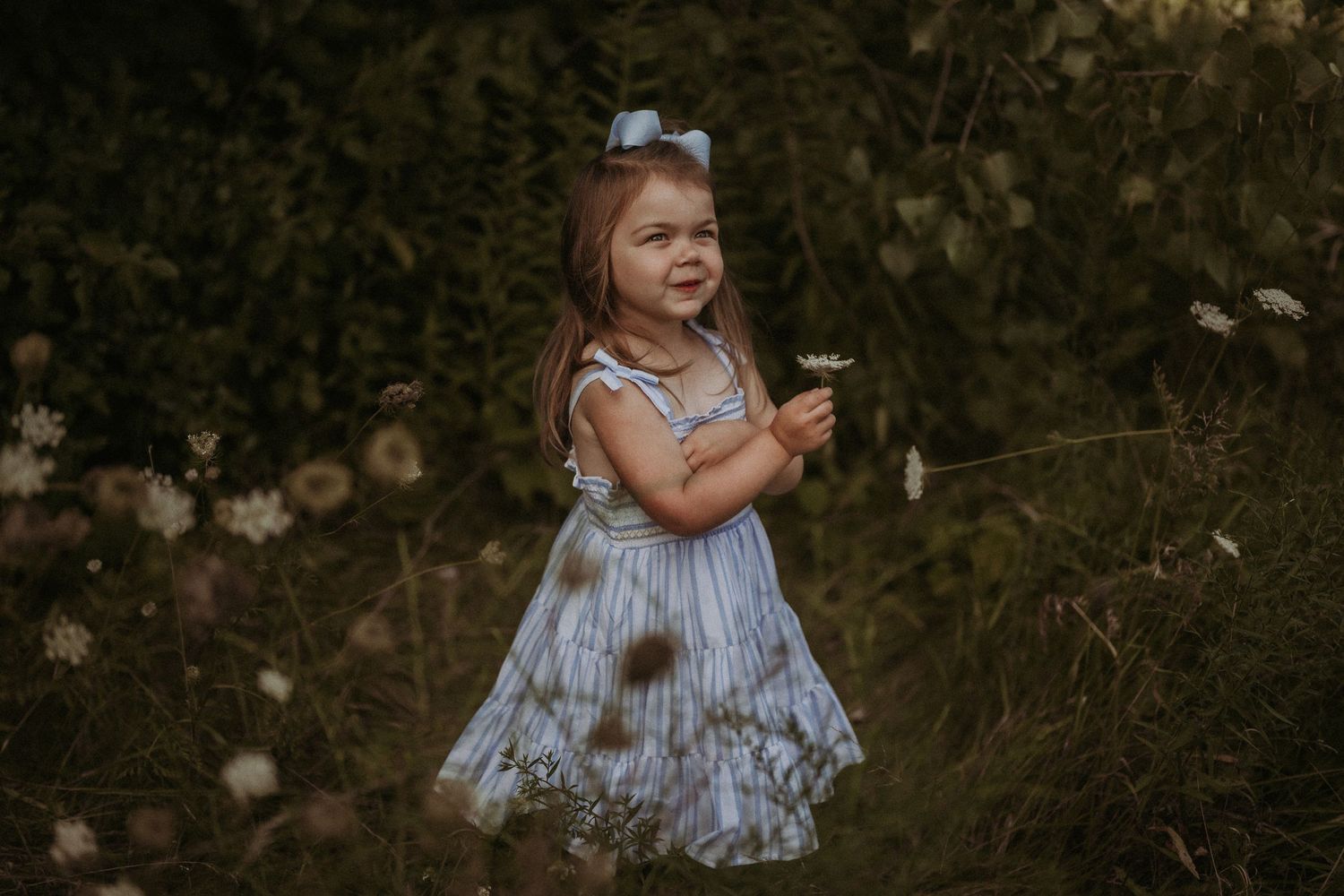 A child in a light dress poses playfully outdoors among wild plants and flowers.