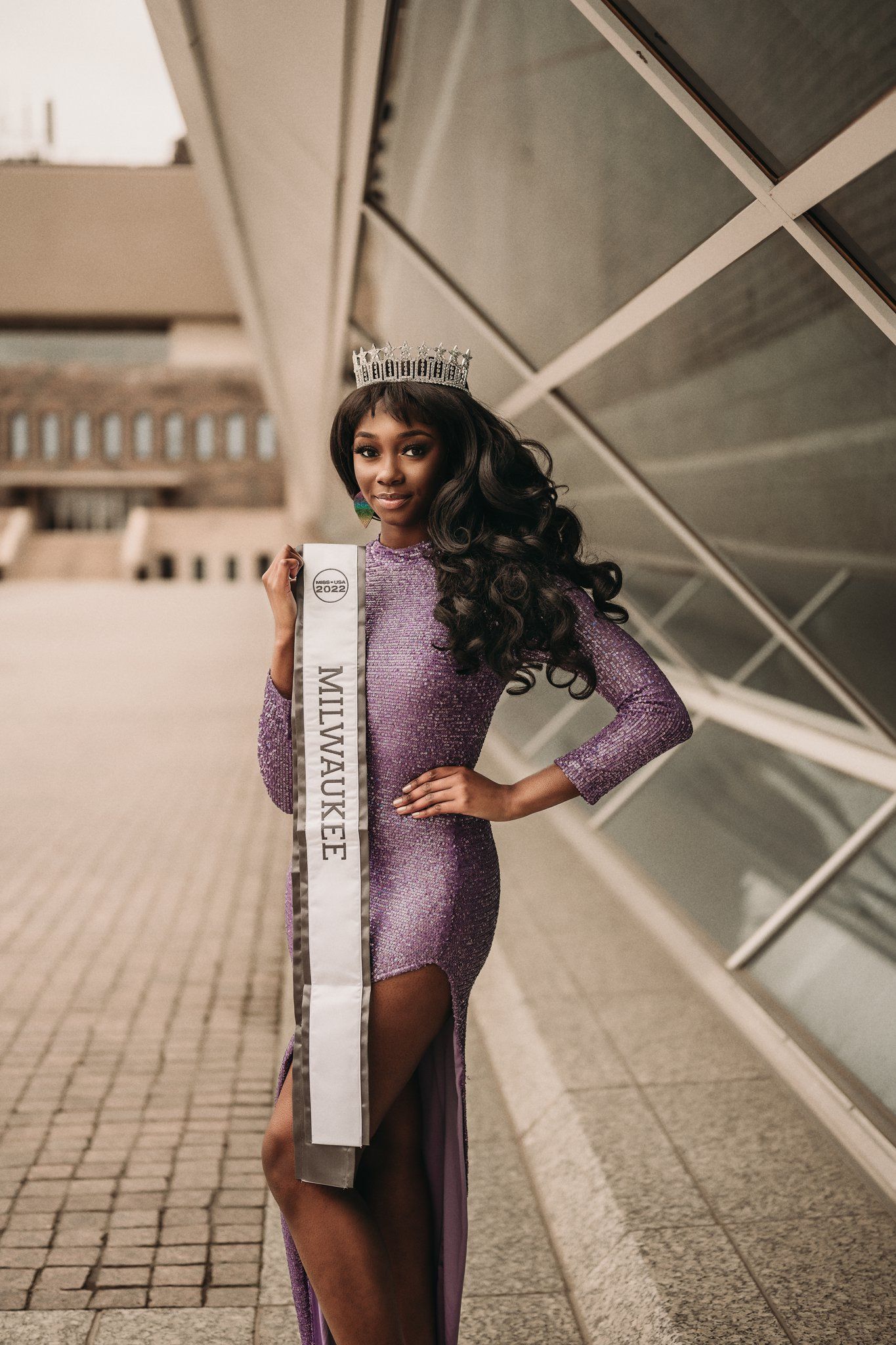 Pageant contestant wearing purple sequin gown and sash poses by modern building.