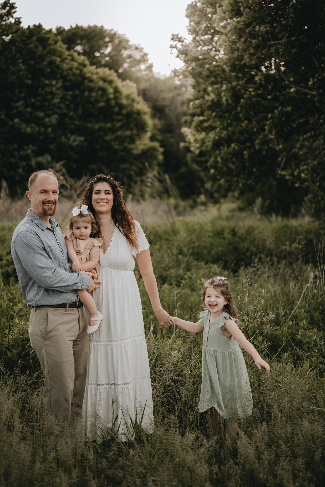 Family portrait in a sunlit meadow with flowing white dress and casual attire creating a dreamy outdoor scene.