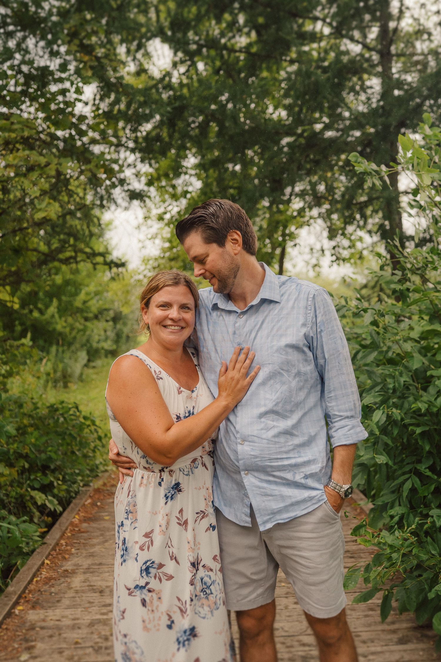 A couple shares an intimate moment while walking on a wooden path surrounded by trees.