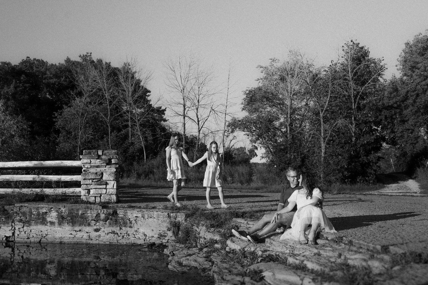 People enjoying a relaxing moment by a rustic pond with trees in black and white photography.
