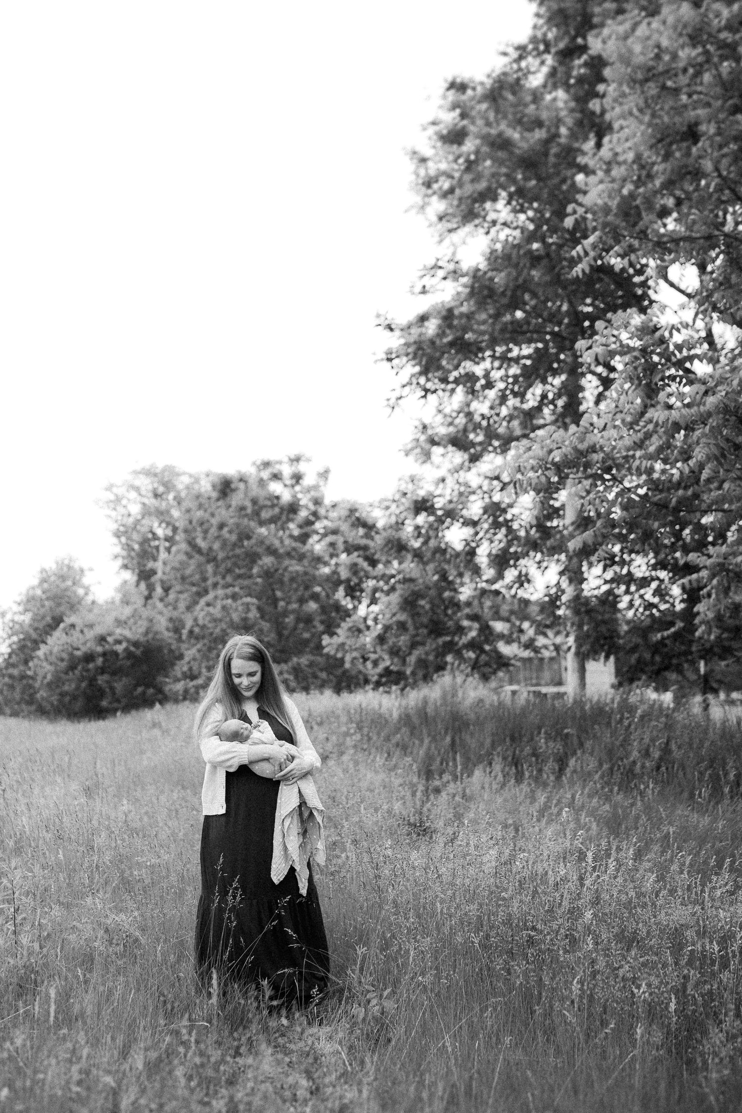 Black and white portrait of a figure standing in a serene meadow with tall trees in the background.