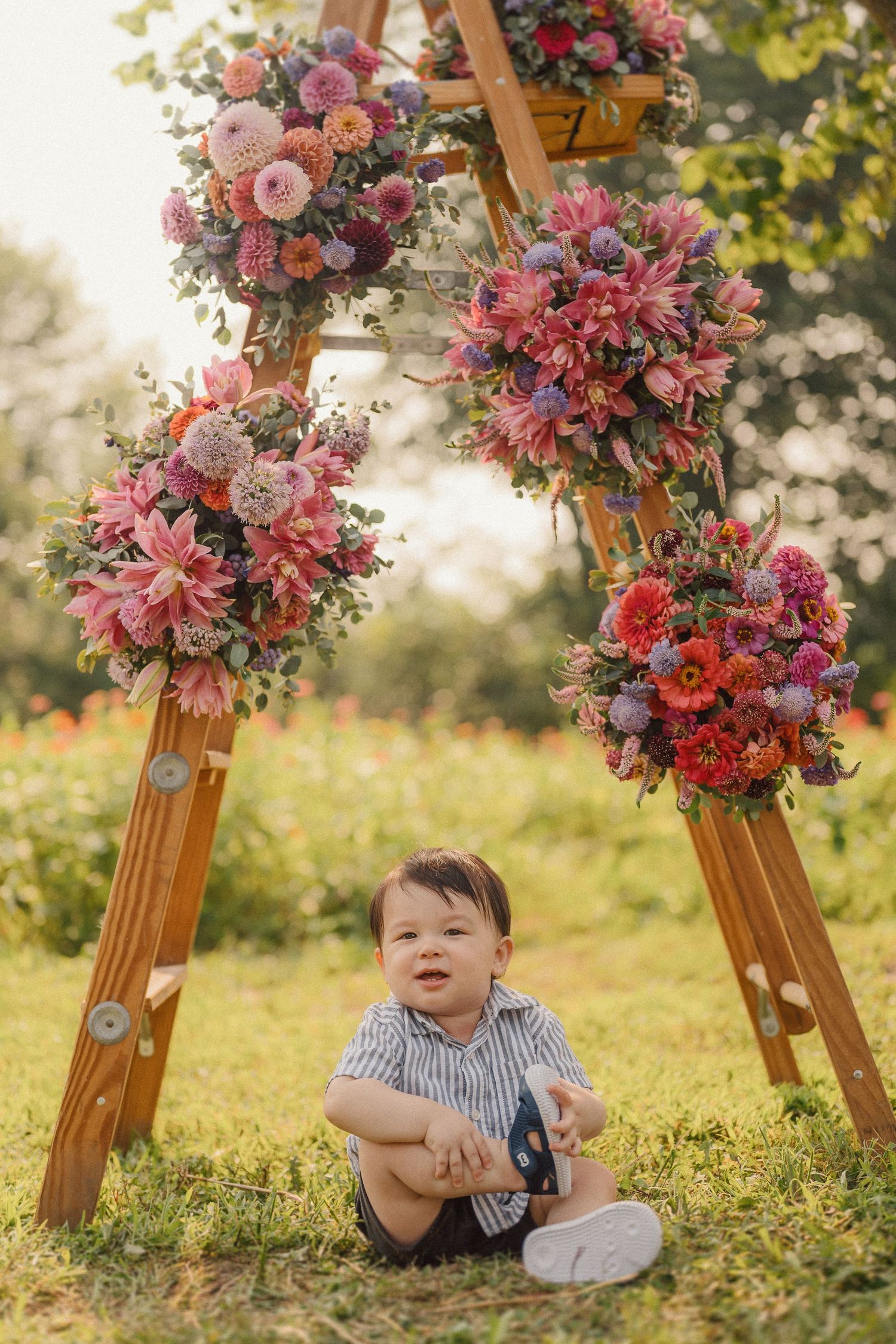 Wooden ladder arch decorated with pink and coral floral arrangements in a sunny meadow setting.