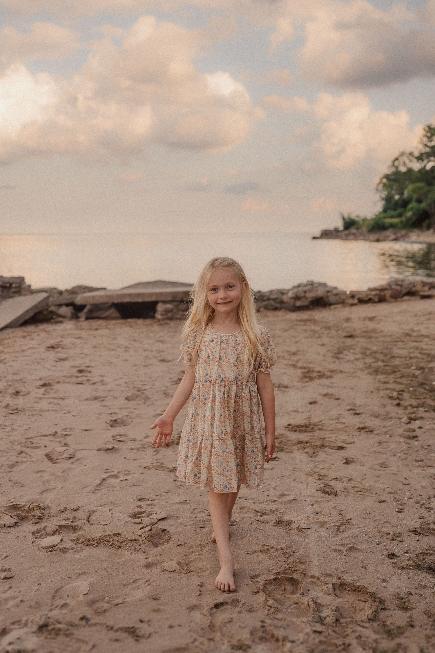 Child in floral dress stands on sandy beach with driftwood and trees under cloudy sky at sunset.