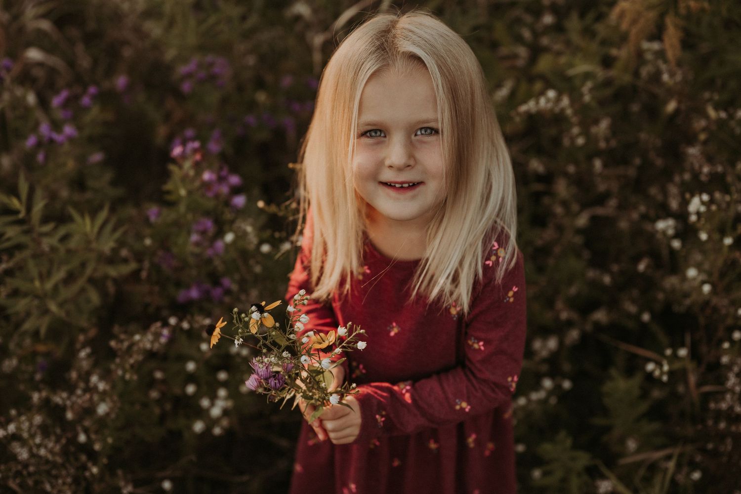 A burgundy dress-clad figure holds wildflowers while standing in a garden during golden hour.