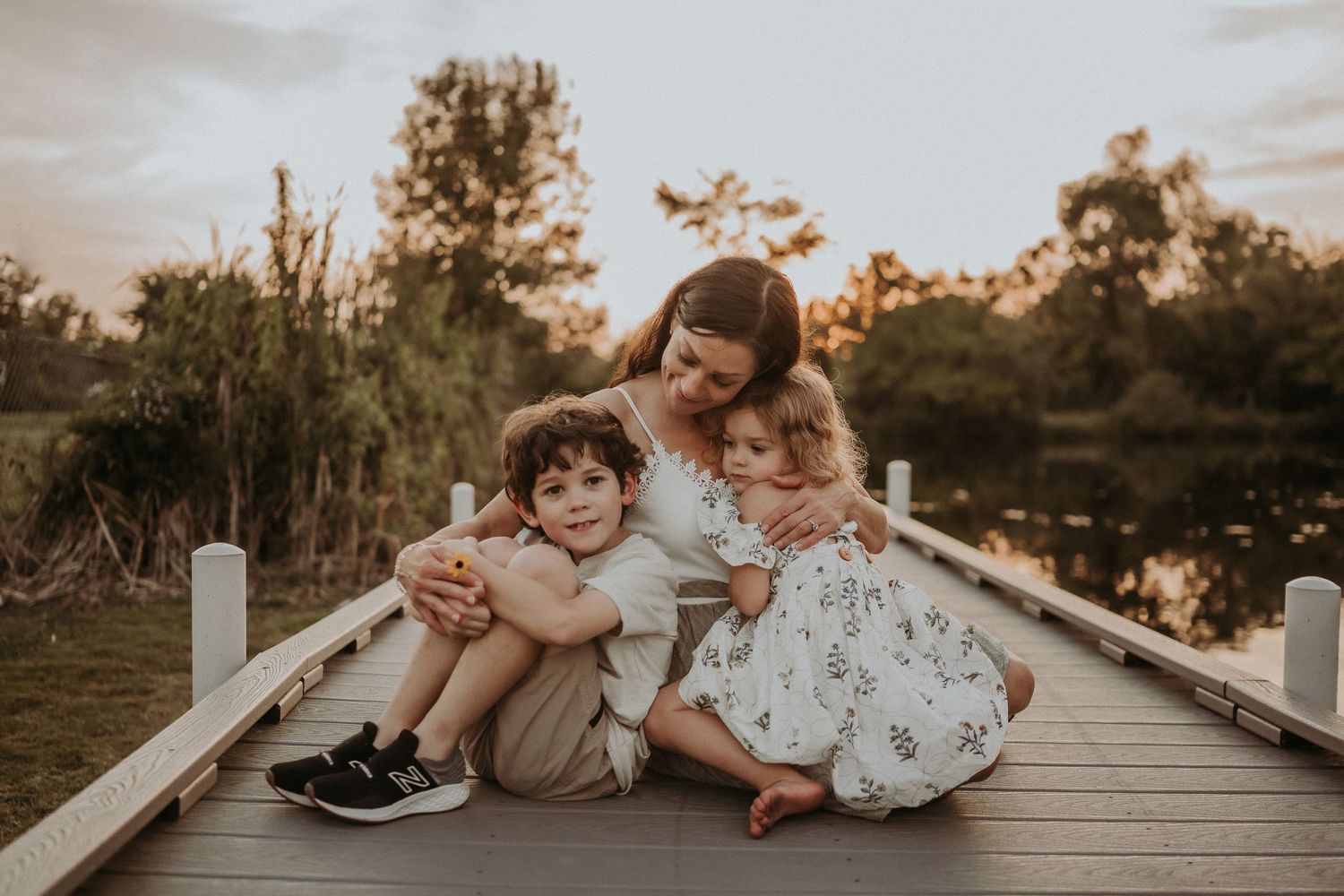 Three people share an intimate embrace while sitting on a wooden boardwalk at sunset.