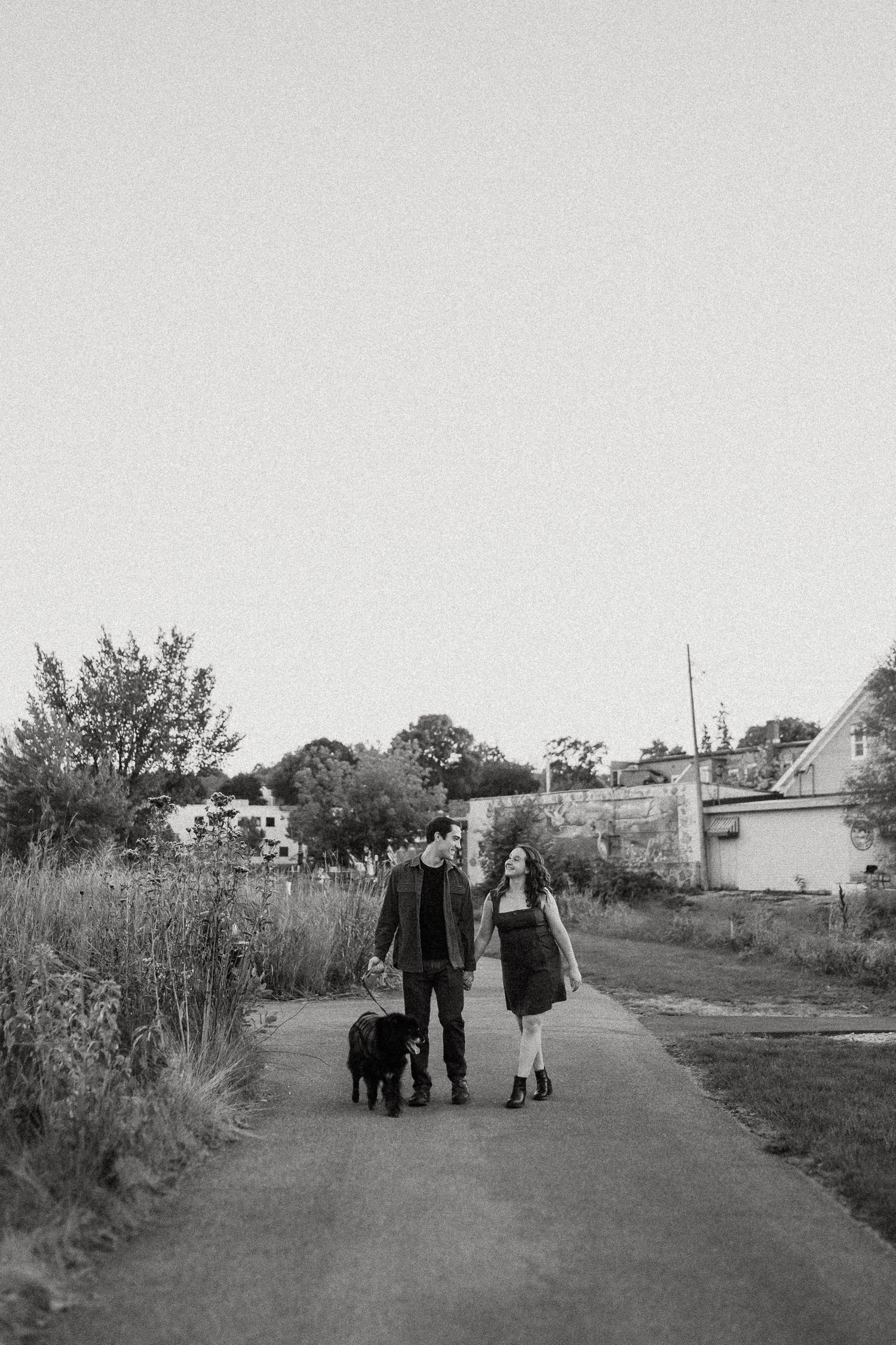 A couple walks their black dog along a countryside path in black and white photography.